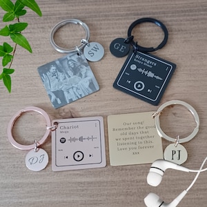 Puede incluir: Cuatro llaveros diferentes con dijes personalizados y etiquetas cuadradas grabadas. Un llavero presenta una foto en blanco y negro de una familia, otro tiene una etiqueta negra con el texto "Strangers Kenya Grace" y un código de barras de Spotify, un tercer llavero tiene una etiqueta de oro rosa con el texto "Chariot Mega" y un código de barras de Spotify, y el último llavero tiene una etiqueta dorada con el texto "¡Nuestra canción! Recuerda los buenos viejos tiempos que pasamos juntos escuchando esto. Te amo para siempre XXX".
