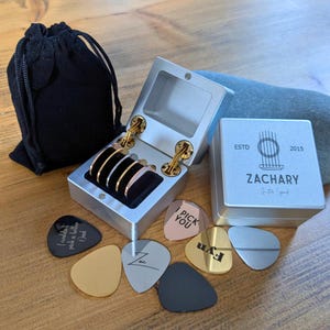 Peut inclure: Une boîte en argent avec un couvercle à charnière contenant un jeu de médiators. La boîte est ouverte et les médiators sont visibles. Il y a aussi plusieurs médiators sur une surface en bois, dont un qui dit "I pick you".