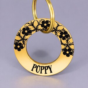 May include: A gold-colored circular keychain with the name "POPPY" in black lettering. The outer edge features a black floral design. A gold ring is attached at the top for easy attachment to keys or a bag.