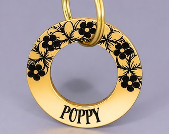 Puede incluir: Un llavero circular dorado con el nombre "POPPY" en letras negras. El borde exterior presenta un diseño floral negro. Un anillo dorado está unido en la parte superior para facilitar la fijación a llaves o una bolsa.