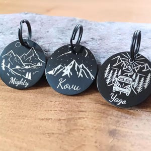 May include: Three black metal pet tags with mountain and nature designs. One tag reads "Mighty", another "Kovu", and the third "Yago". Each tag has a black ring for attachment.