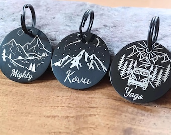Könnte beinhalten: Drei schwarze Metall-Haustieranhänger mit Berg- und Naturmotiven. Ein Anhänger trägt die Aufschrift "Mighty", ein anderer "Kovu" und der dritte "Yago". Jeder Anhänger hat einen schwarzen Ring zur Befestigung.