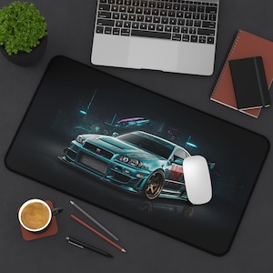 Pode incluir: Tapete de mesa preto com uma ilustração de um carro desportivo turquesa com jantes douradas. O carro está situado num cenário urbano escuro e futurista. Um rato branco, um portátil e artigos de papelaria também são visíveis.