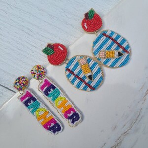 May include: A pair of colorful beaded earrings with a teacher theme. One pair features a red apple and a blue and white striped design with a yellow pencil. The other pair features the word "TEACHER" in rainbow colors.