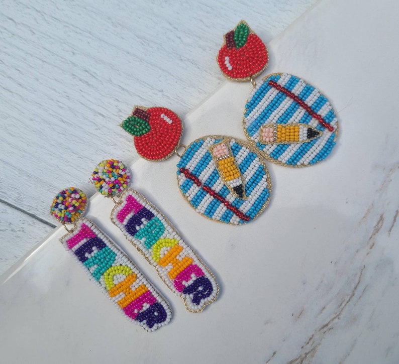 May include: A pair of colorful beaded earrings with a teacher theme. One pair features a red apple and a blue and white striped design with a yellow pencil. The other pair features the word "TEACHER" in rainbow colors.