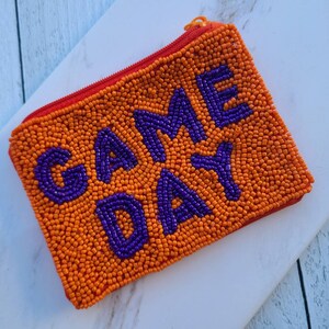 Pu&ograve; includere: Porta monete in perle arancioni con la scritta "GAME DAY" in perle viola.