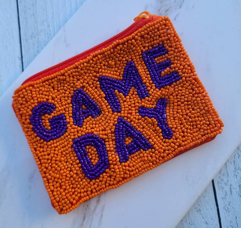 Pu&ograve; includere: Porta monete in perle arancioni con la scritta "GAME DAY" in perle viola.