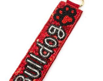 Puede incluir: Un llavero rojo con lentejuelas con la palabra "Bulldog" en cuentas negras y plateadas. El llavero presenta un diseño de huella de pata y un cierre dorado.