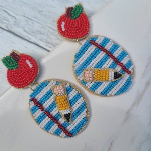 May include: A pair of beaded earrings with a red apple and a blue and white striped design with a yellow pencil.