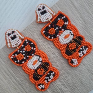 Puede incluir: Un par de pendientes de cuentas naranja y negro con un amuleto de fantasma blanco colgando de cada pendiente.