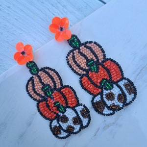 May include: A pair of dangle earrings with a stack of orange and white beaded pumpkins. The pumpkins are decorated with green leaves and black beads. The earrings are topped with a bright orange flower.