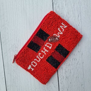 Pu&ograve; includere: Portafoglio rosso con cerniera in perline con la scritta "TOUCHDOWN" in bianco e un'immagine di un pallone da calcio.