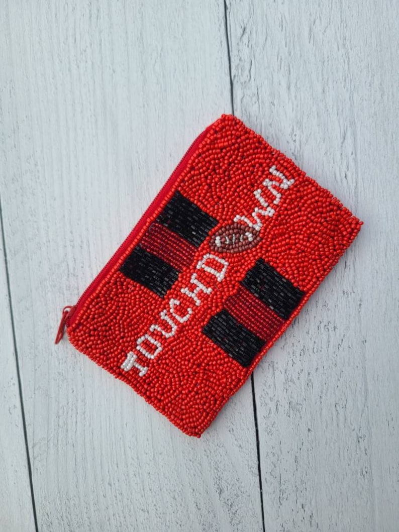 Pu&ograve; includere: Portafoglio rosso con cerniera in perline con la scritta "TOUCHDOWN" in bianco e un'immagine di un pallone da calcio.