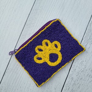 Pu&ograve; includere: Un piccolo porta monete di perline viola con un design a forma di zampa gialla.