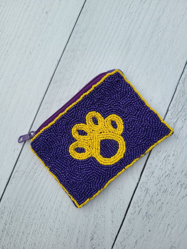 Pu&ograve; includere: Un piccolo porta monete di perline viola con un design a forma di zampa gialla.
