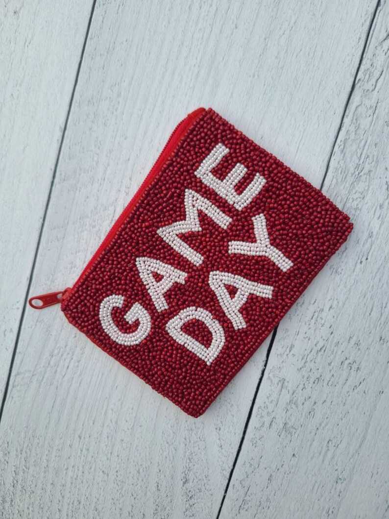 Pu&ograve; includere: Portafoglio rosso con cerniera a forma di perline con le parole "GAME DAY" in perline bianche.