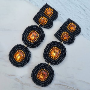 Könnte beinhalten: Ein Paar schwarze Perlenohrringe mit orangefarbenen Edelstein-Akzenten. Die Ohrringe sind in Form eines Buchstabens "B" gestaltet und haben drei runde Perlen mit Edelsteinen in der Mitte.