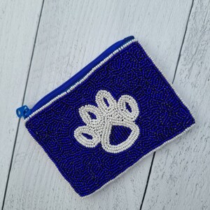 Pu&ograve; includere: Un piccolo porta monete blu perlato con un design a forma di zampa bianca. Il porta monete ha una chiusura a zip.