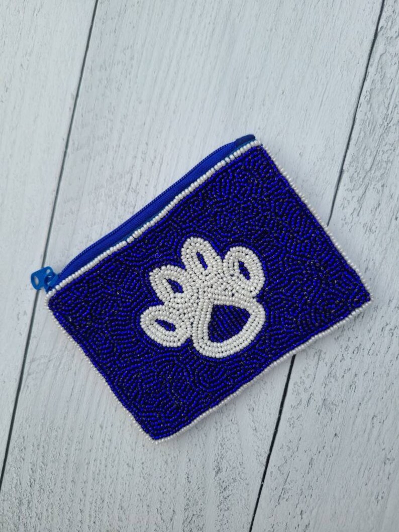 Pu&ograve; includere: Un piccolo porta monete blu perlato con un design a forma di zampa bianca. Il porta monete ha una chiusura a zip.