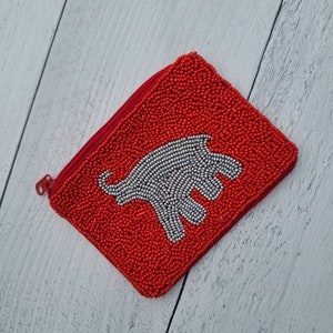 Pu&ograve; includere: Un porta monete rosso di perline con un design di elefante bianco di perline.