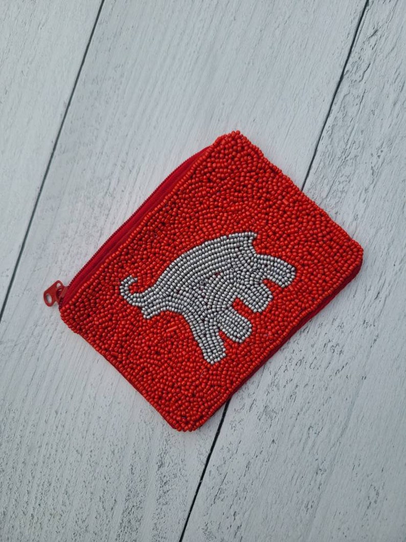 Pu&ograve; includere: Un porta monete rosso di perline con un design di elefante bianco di perline.