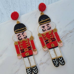 May include: A pair of beaded nutcracker earrings. The earrings are red, white, and black with gold accents. The nutcrackers are wearing red coats with gold buttons and black hats.
