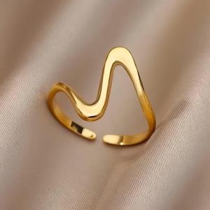 Puede incluir: Un anillo de color dorado con un diseño ondulado abstracto. El anillo es abierto y ajustable.