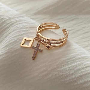 Puede incluir: Anillo dorado con tres bandas y un dije colgante con una cruz y un cuadrado con diamantes de imitación.
