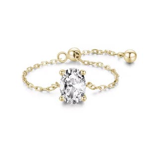 Puede incluir: Un anillo de cadena de oro con una gran piedra preciosa ovalada transparente. La cadena es ajustable y tiene una pequeña perla dorada en cada extremo.