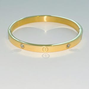 Puede incluir: Un brazalete de oro con acabado pulido y pequeñas piedras transparentes engastadas en la banda. El brazalete tiene un diseño simple y elegante.