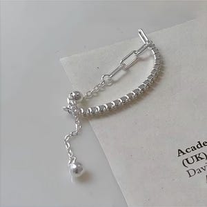 Puede incluir: Un brazalete de cadena de plata con un dije colgante. El brazalete está hecho de pequeños eslabones cuadrados y tiene una cadena delicada con un pequeño dije de bola.