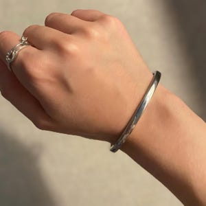 Puede incluir: Se muestra una pulsera de plata y un anillo de plata. La pulsera es una banda simple y pulida. El anillo tiene un diseño delicado. Las joyas se muestran en la mano de una persona.