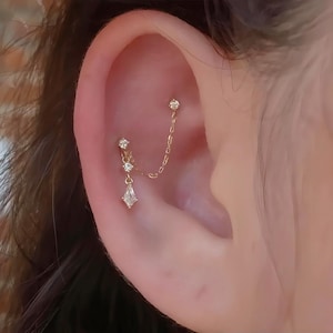 Puede incluir: Primer plano de la oreja de una persona con una cadena de oro y pendientes de diamantes. La cadena está unida a un pendiente de diamante en la parte superior de la oreja y tiene tres diamantes más pequeños colgando de ella. La cadena termina en un colgante de diamante más grande.