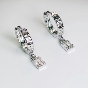 Puede incluir: Un par de pendientes de aro de plata con un colgante rectangular adornado con piedras de circonita cúbica transparentes.