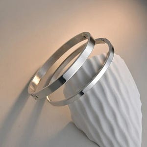 Puede incluir: Dos pulseras de brazalete plateadas con bisagras. Las pulseras tienen una superficie plana y pulida y un diseño simple y moderno. Se exhiben sobre una escultura abstracta blanca y texturizada, creando una estética minimalista. Las pulseras son reflectantes.