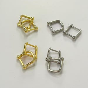 May include: Two pairs of gold and silver hoop earrings with a square shape and a ball detail at each corner. The earrings are stacked on top of each other.