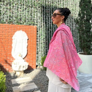May include: A woman wearing sunglasses and a pink and white marbled scarf. The scarf has a flowing, draped design. The woman is standing in front of a brick wall with a white statue.