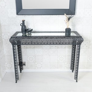 May include: Black metal console table with intricate cut-out designs and a mirrored top. The table has two legs on each side and is decorated with a small vase and a bowl.