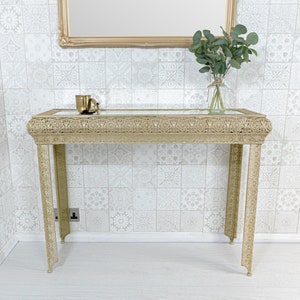 May include: A gold metal console table with a glass top and intricate filigree detailing. The table has a rectangular top and four legs. The table is in front of a white tiled wall.