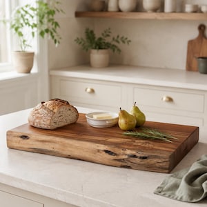 May include: A rectangular wooden chopping board with a natural live edge, featuring a loaf of crusty bread, a small white ceramic dish containing a square of butter, two green pears, and a sprig of fresh rosemary arranged on its surface.