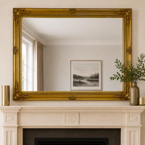 May include: A large rectangular wall mirror featuring a wide, ornate gold frame with intricate corner and centre detailing. The mirror surface reflects a room interior, including a framed landscape print, a window, and light-coloured walls.