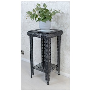 May include: Black metal plant stand with a decorative, intricate design. The stand has a glass top and a lower shelf. A potted plant sits on the top shelf.