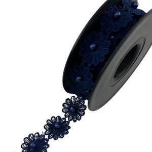 Könnte beinhalten: Eine Spule mit dunkelblauem Spitzenband mit einem sich wiederholenden Blumenmuster. Jedes einzelne Blumenmotiv ist in der Mitte mit einer kleinen, runden, passenden dunkelblauen Perle verziert. Die Spitze ist auf eine schwarze Kunststoffspule gewickelt.