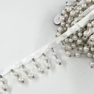 May include: White satin ribbon with dangling faux pearl beads. The beads are strung on silver-colored wire and hang in small clusters. The ribbon is on a white background.
