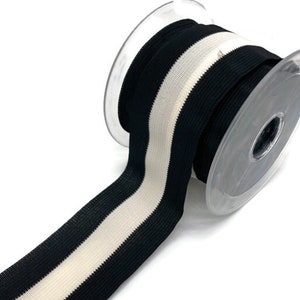 Könnte beinhalten: Eine Rolle schwarz-weiß gestreiftes elastisches Band. Das Band ist auf einer transparenten Kunststoffspule aufgewickelt, wobei die Streifen in Längsrichtung verlaufen. Das elastische Material ist sichtbar und das Band ist für Näh- oder Bastelprojekte geeignet.