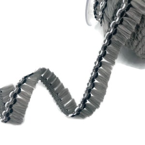 May include: Grey fringe trim with black and white accents. The trim has a tassel-like appearance and is made of a soft, fuzzy material.