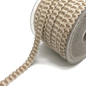 May include: A spool of decorative trim featuring a looped, scalloped design. The trim is a neutral beige color with a lighter beige border, perfect for adding a textured accent to craft projects. The trim is wound on a clear plastic spool.