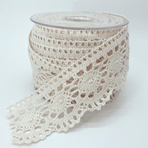 May include: A spool of off-white crochet lace trim featuring a repeating circular floral pattern with scalloped edges. The lace is wound tightly around a clear plastic cylindrical spool, showcasing the intricate woven texture of the cotton thread.