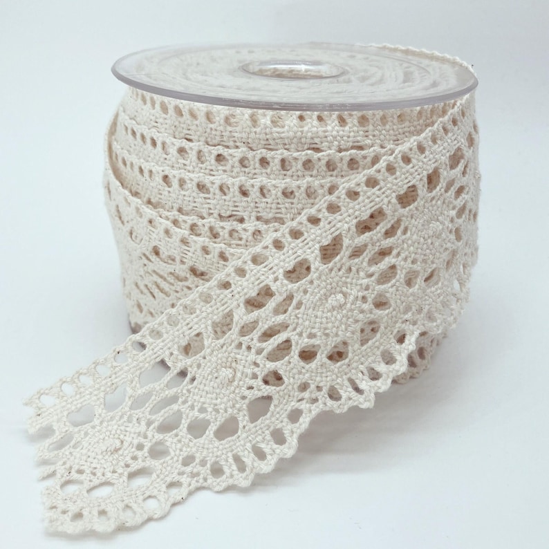May include: A spool of off-white crochet lace trim featuring a repeating circular floral pattern with scalloped edges. The lace is wound tightly around a clear plastic cylindrical spool, showcasing the intricate woven texture of the cotton thread.