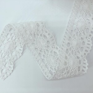 May include: A white lace trim featuring a repeating circular pattern with intricate openwork details. The fabric has a scalloped edge and a continuous design, showcasing a delicate, woven texture throughout the entire length of the material.